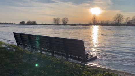 waves coming in on the city boulevard riverbed of a flooded river ijssel reaching a bench during rising and high water levels at sunset with vast floodplain landscape trees under water on the horizon