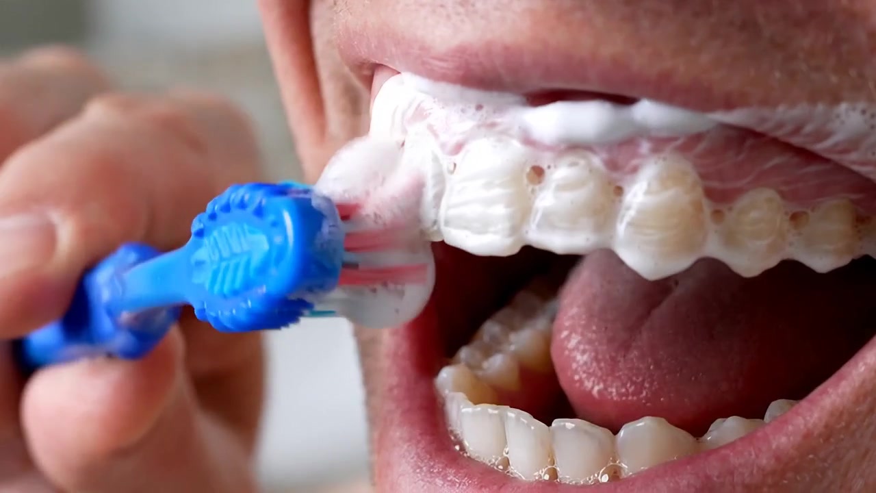 Primo piano di uno spazzolino blu che pulisce denti molto puliti con schiuma attorno alla bocca di un uomo con barba incolta