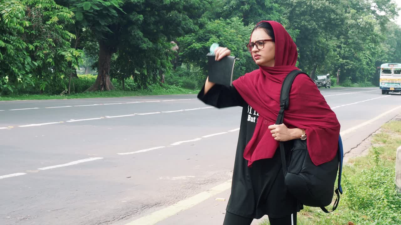 Beautiful serious young Afghan woman in hijab holding file and bag in hand and standing on street near road car passing in the background, charming female in black t shirt