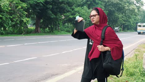 beautiful serious young afghan woman in hijab holding file and bag in hand and standing on street near road car passing in the background, charming female in black t shirt