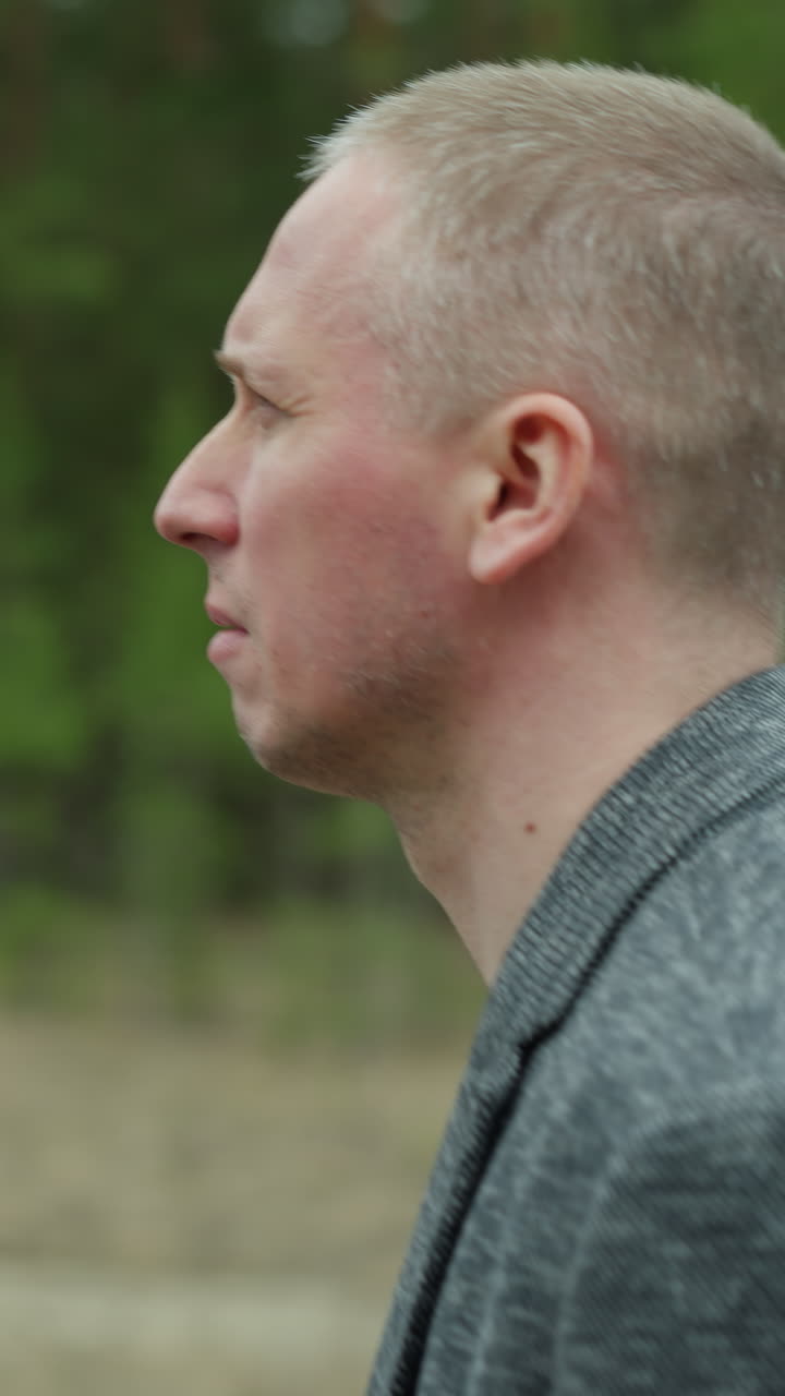 A close-up side profile of a middle-aged man with short hair, wearing a grey suit jacket and a black shirt, walking outdoors with a blurred forest background