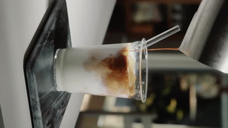 vertical video home barista making iced latte pouring espresso shot into glass drinking cup containing milk with glass straw in slow motion close up