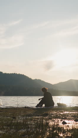 a la señora le gusta lanzar piedras al río al atardecer. la mujer descansa en un picnic sentada en la orilla del río con una vista impresionante de las montañas. cálida noche de otoño