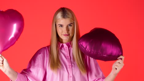 Pretty-Blonde-Woman-Holding-Waving-Purple-Heart-Shaped-Balloons-Against-Red-Background,-Colorful-Studio-Shot