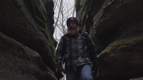 a hiker wearing a backpack and a flannel shirt enter a rocky crevice on a cold winter day before turning to reveal a gray sky and barren trees