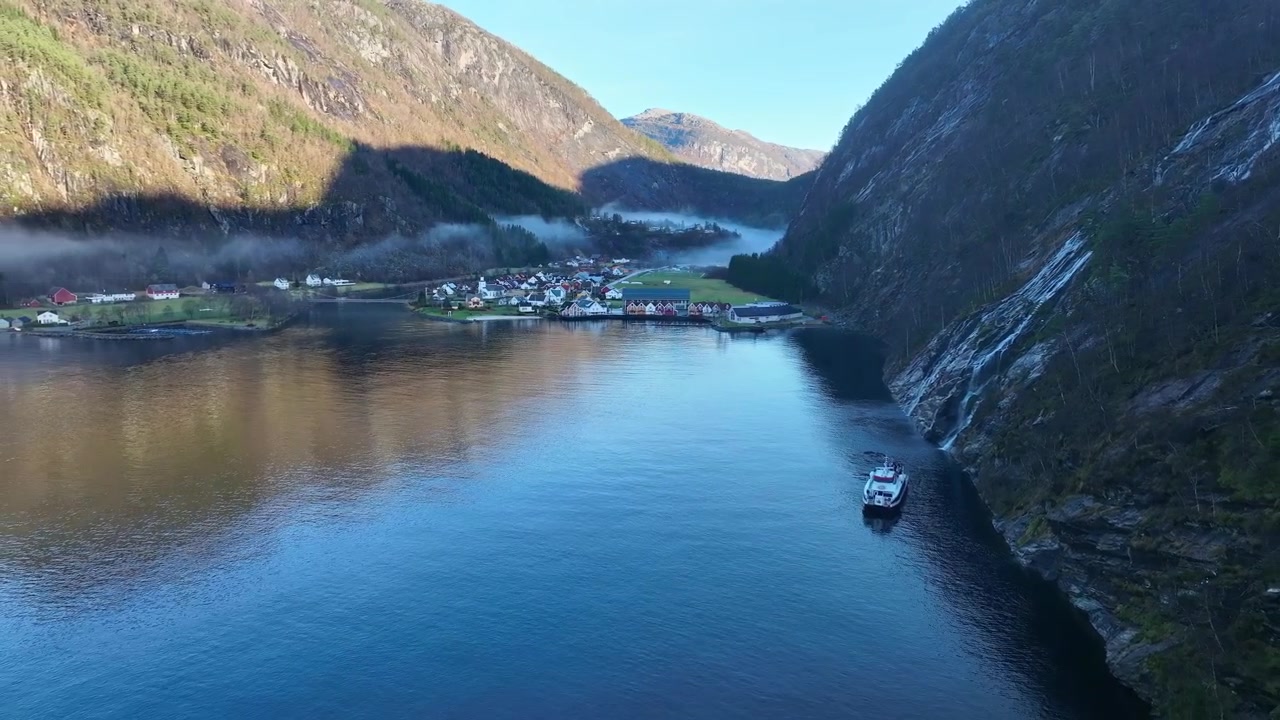 Luftaufnahme, die sich rückwärts über einen Fjord in Mo, Norwegen, bewegt und dabei das Stadtzentrum, Berge und einen Touristenkatamaran nahe eines Wasserfalls enthüllt
