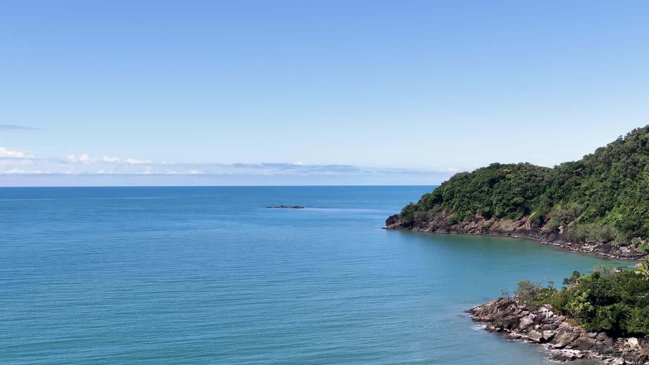 Smooth aerial pan reveals lush rainforest headland meeting calm blue sea under bright daylight