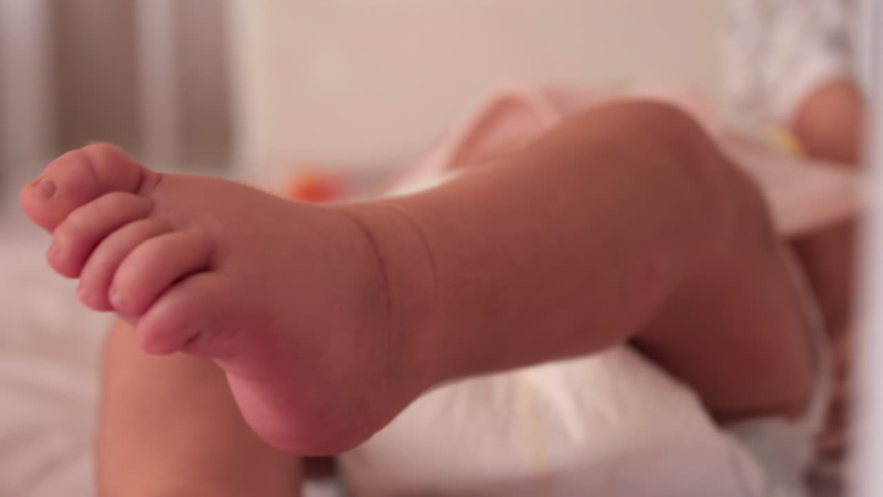 Close up of a baby's legs and diaper as the infant rests in a crib, seen through the crib bars