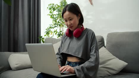 young asian woman typing on laptop while wearing casual clothes, working on comfort sofa in living room at home