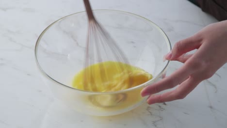 a hand whisks eggs in a transparent glass bowl on a bright marble countertop under soft natural light.