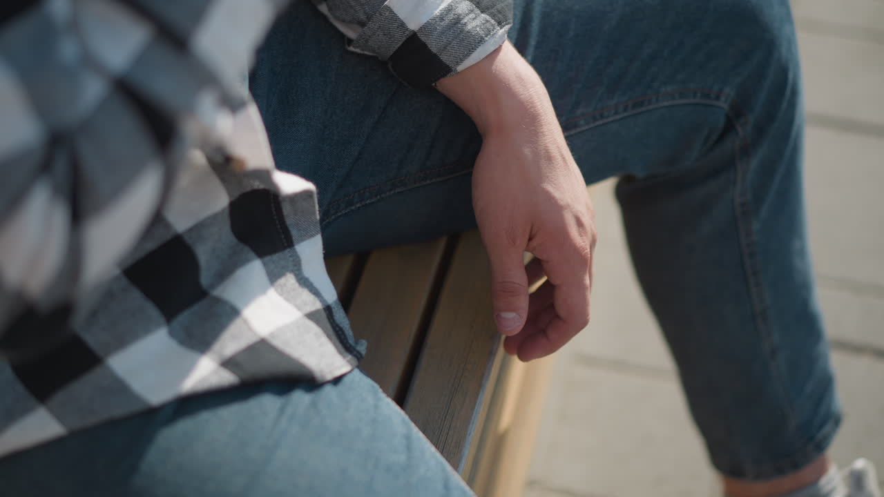 close up of fair skinned student wearing checkered shirt and jeans sitting on wooden bench with relaxed hand after taking red pill medicine on bright sunny day outdoors with pavement background