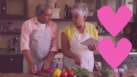 two pink heart icons against african american senior couple wearing aprons cooking together at home