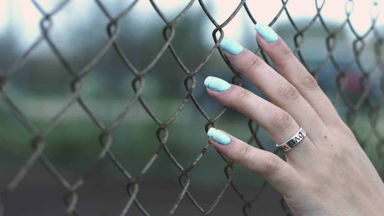 Mano femminile con anello e manicure blu che tocca la rete di una recinzione mentre cammina per strada.