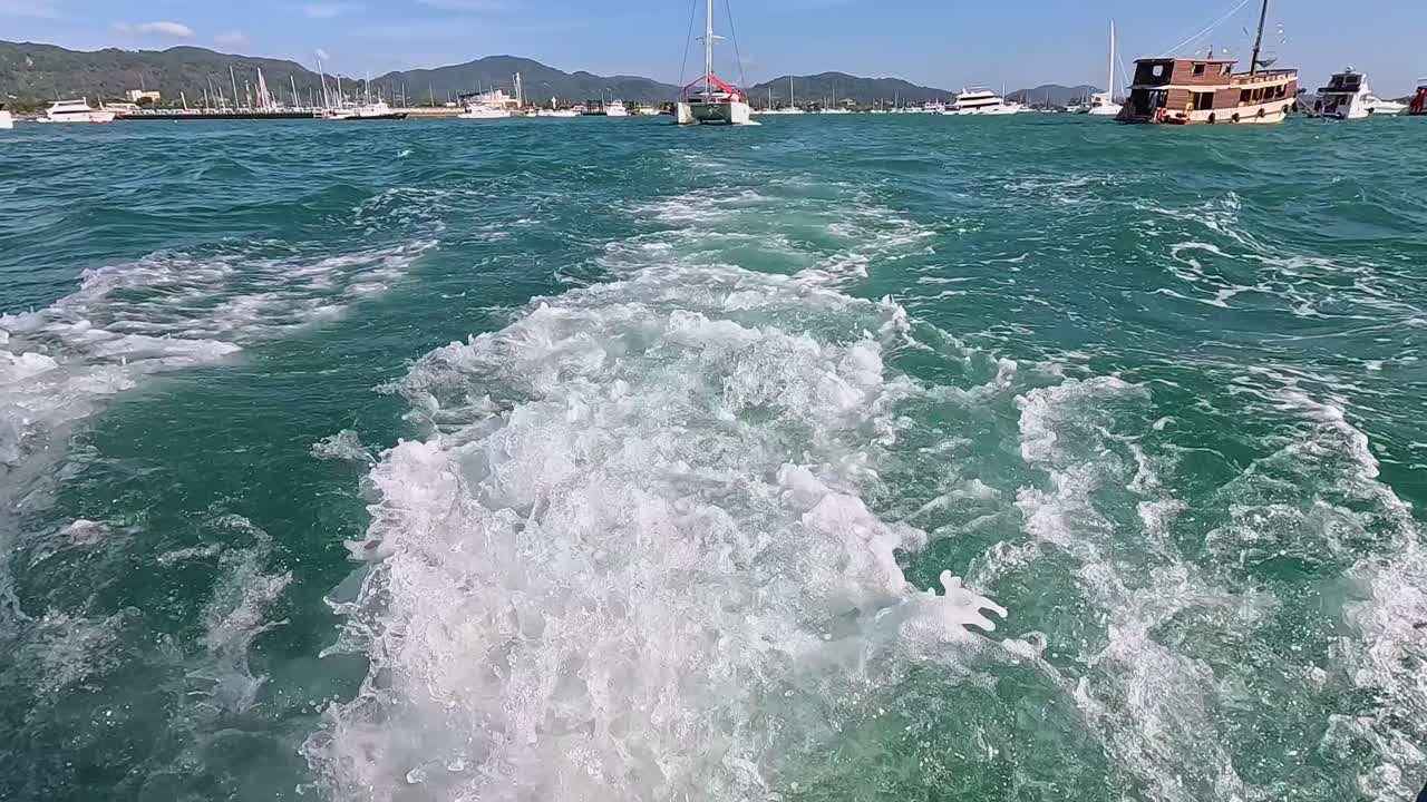 A catamaran sails through Phuket's turquoise waters, leaving a foamy wake under clear skies and bright sunlight