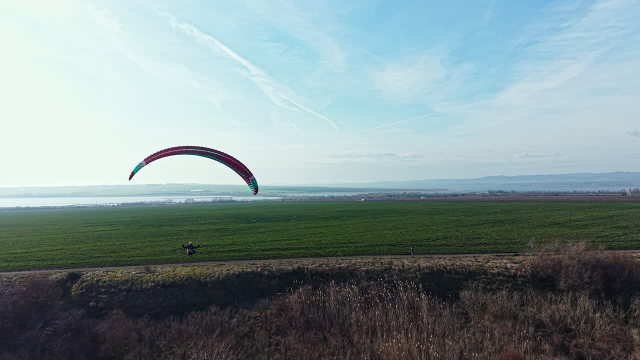 Paraglider flyver over grøn mark med bakker i baggrunden på en klar dag i åbent landskab.