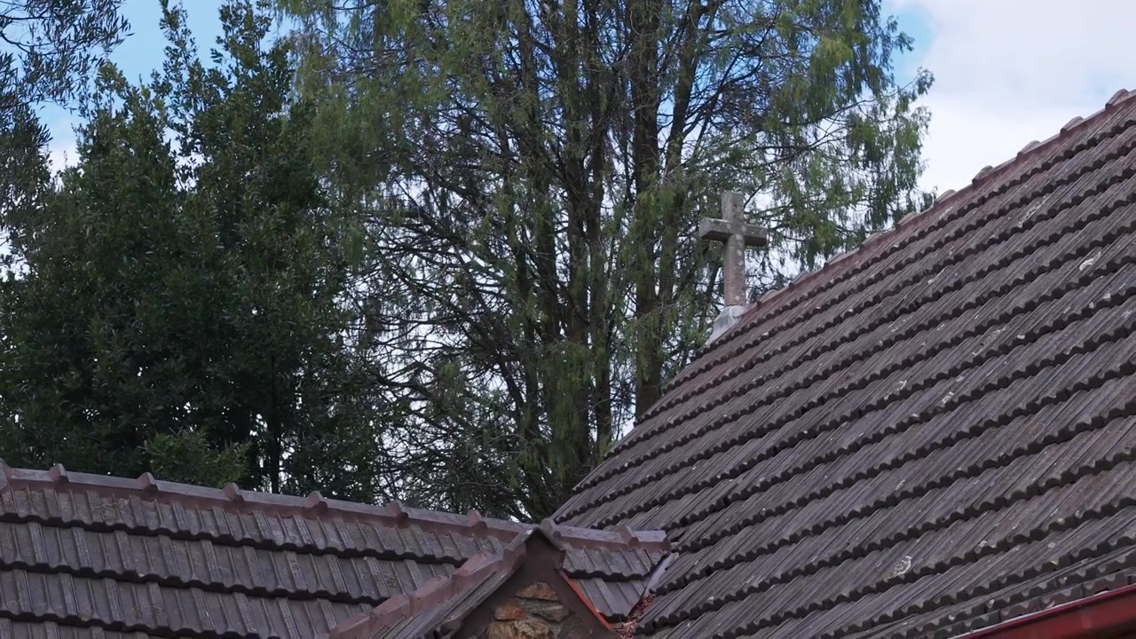 Bidikan lebar gereja batu tua di Olinda, Melbourne, menunjukkan atap genteng, salib, dan pohon-pohon hijau. Cahaya siang menonjolkan tekstur pedesaan, latar alami, dan detail arsitektur yang tenang.