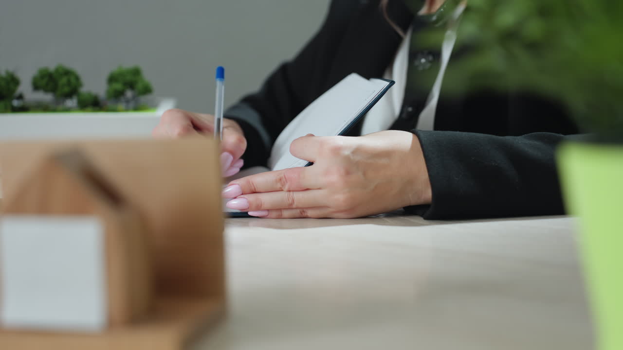 close up of woman with polished pink nails writing in notebook beside house miniature model on desk in bright office space with natural light and blurred green plant in foreground