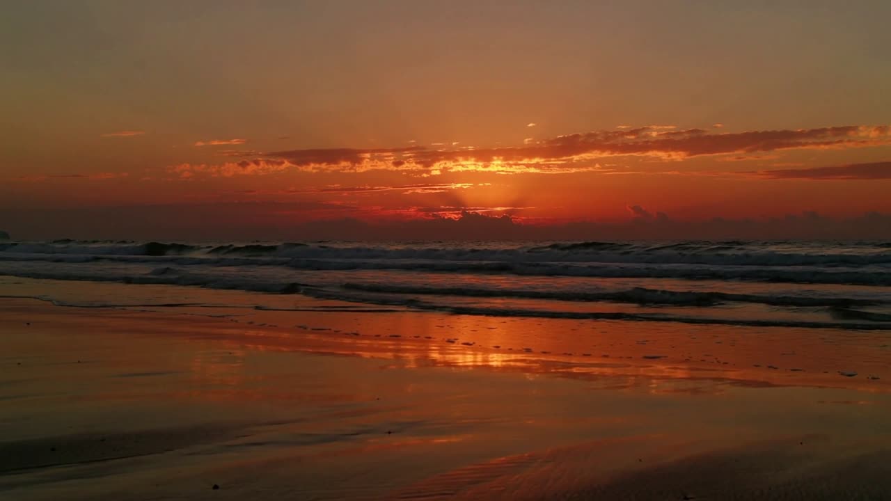 Casting Sun at horizon sending glow across wet sand as cloud band shifting and waves lapping