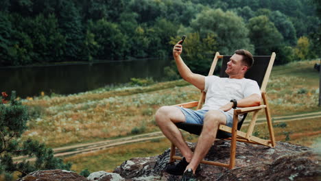 cheerful smiling caucasian man having chat online on his phone. man sits in the folding chair in the nature.
