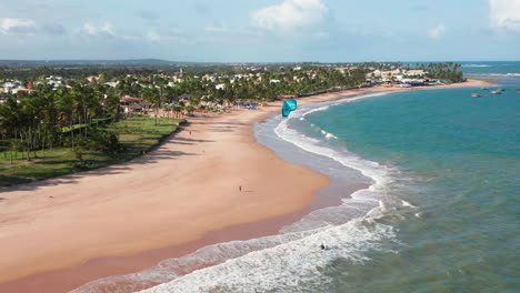 Aerial-view-of-the-beach,-waves-and-a-large-green-area-with-palm-trees,-a-person-practing-kite-surf-and-the-city-at-background,-Guarajuba,-Bahia,-Brazil