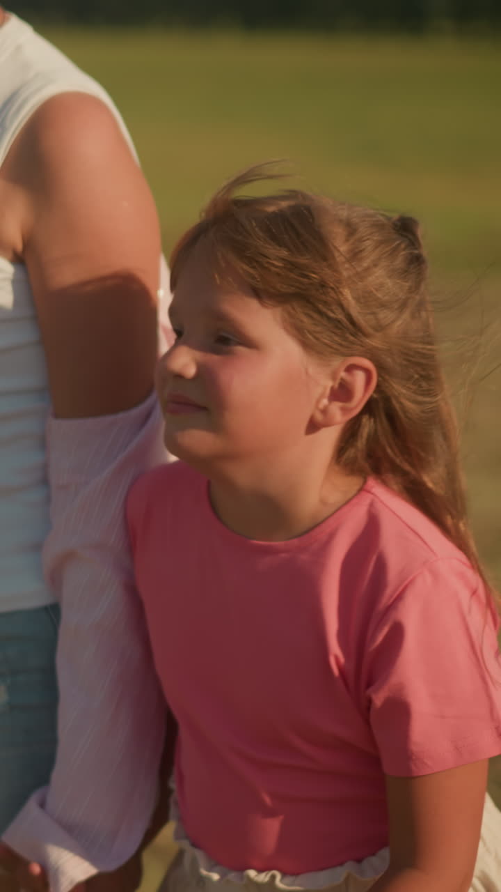 작은 소녀와 이모가 빛의 시골에서 손을 잡고 걸어가는 측면 뷰, 이모는 꽃을 들고, 둘 다 따뜻하게 미소 짓고, 흐릿한 녹색 풍경으로 열린 에서 여름 날을 즐기고 있습니다.