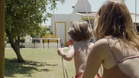 back view of a caucasian mother in swimsuit swinging boy on wooden swing in the backyard