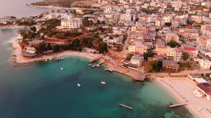 Vista de drone na Albânia voando sobre água azul cristalina e praia de areia branca ao pôr do sol, com hotéis em Ksamil
