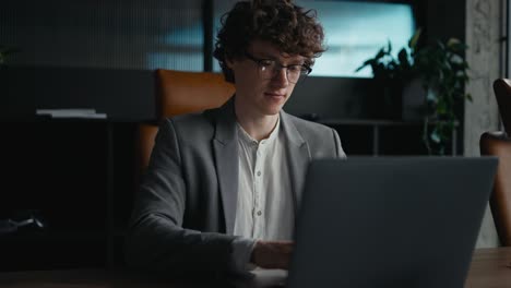 a young guy with curly hair in glasses and a gray jacket works at a laptop sitting at a table in the office