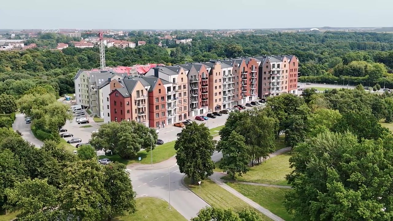 Farverige mellemhøje lejlighedsblokke buer omkring et parkeringsområde ved siden af grønne bygader og åbne græsplæner på en klar sommerdag