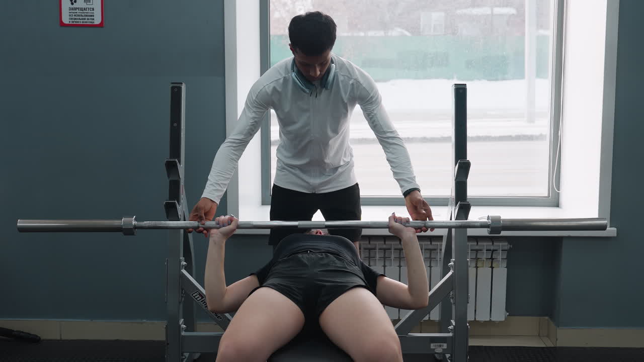 female client lying on bench press receiving spotting assistance from trainer under barbell lift with urban traffic visible through window with modern racks and sunlight