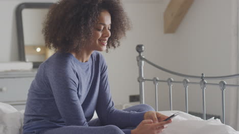 Smiling-Young-Woman-Sitting-On-Bed-With-Mobile-Phone-Wearing-Pyjamas