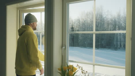 man in yellow hoodie and beanie looking out at snowy landscape from a cozy interior and leaving