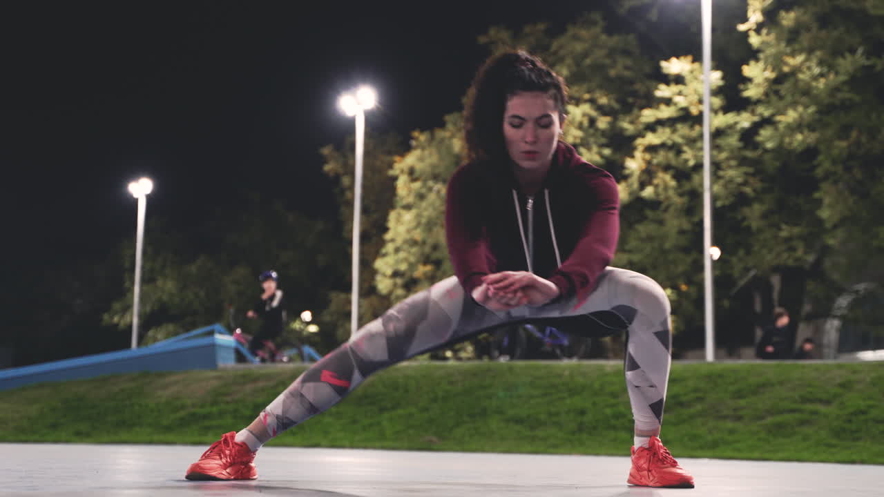 chica rizada deportiva estirando las piernas y haciendo estocadas laterales en el parque por la noche