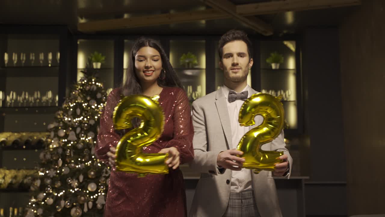 Woman and man wearing elegant clothes holding balloons with the numbers 22 while dancing, kissing and hugging each other in new year's party