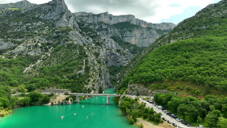 Gorges-du-verdon-with-turquoise-river-and-bridge-surrounded-by-lush-green-mountains,-aerial-view