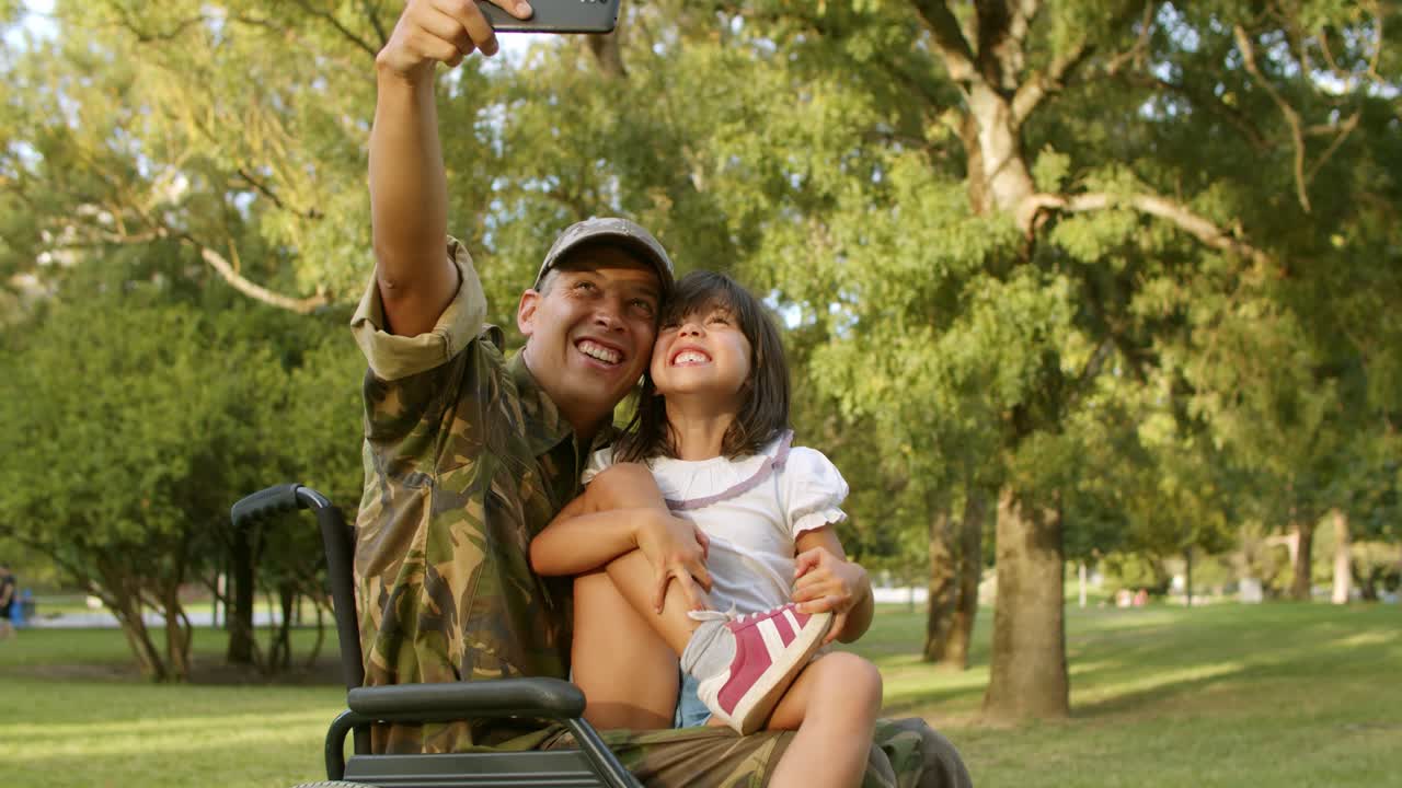 padre militar feliz en silla de ruedas e hija tomando selfie