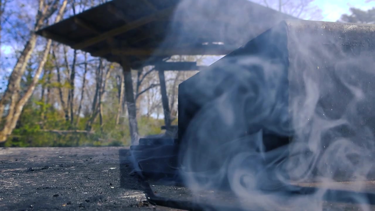 Opstijgende rook van een openbare barbecue op een picknickplaats in het bos, met een houten prieel zichtbaar op de achtergrond op een zonnige dag. Een close-up van de smeulende houtskool.