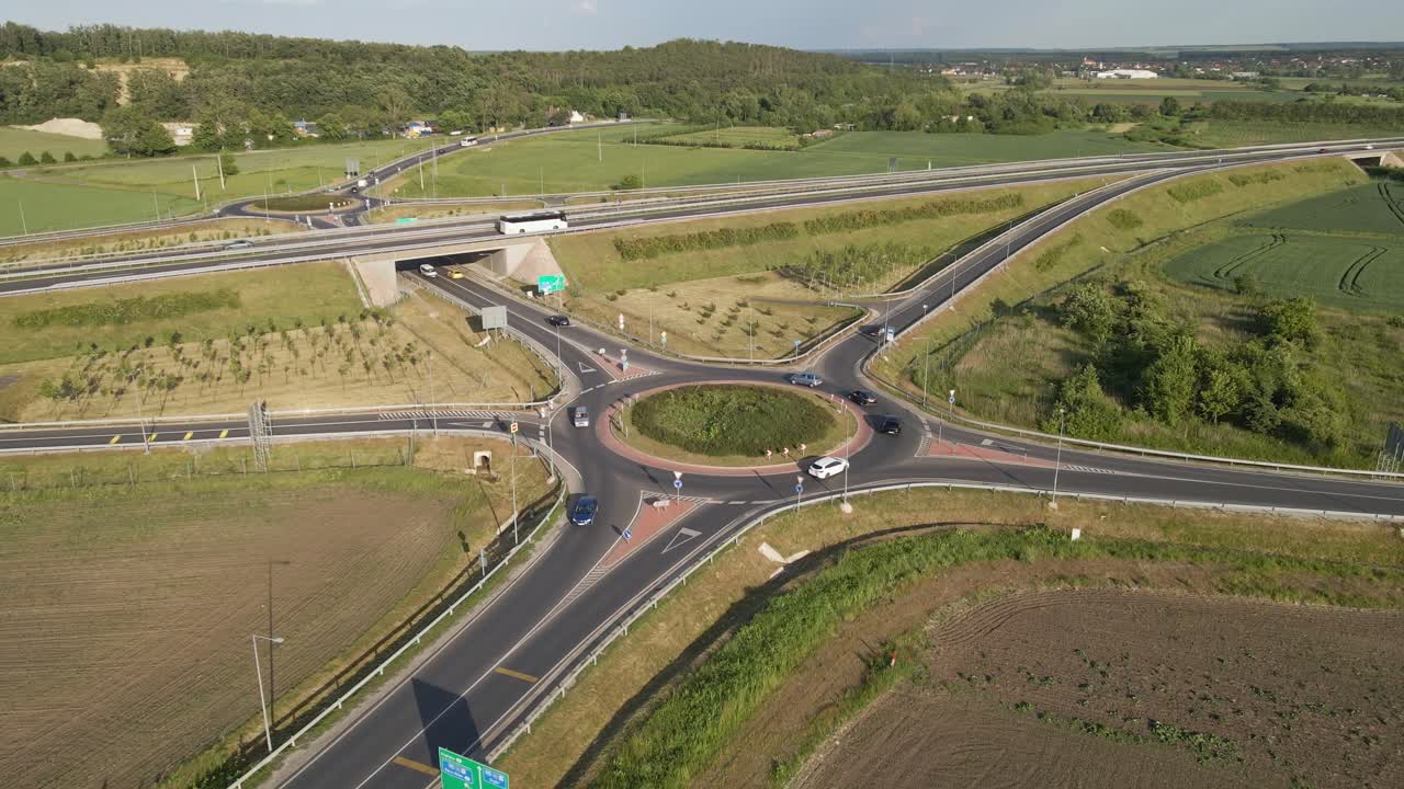 Aerial view of a modern roundabout connecting to a highway interchange in the countryside. The video shows cars navigating the roundabout, surrounded by green fields and farmland under a clear sky.