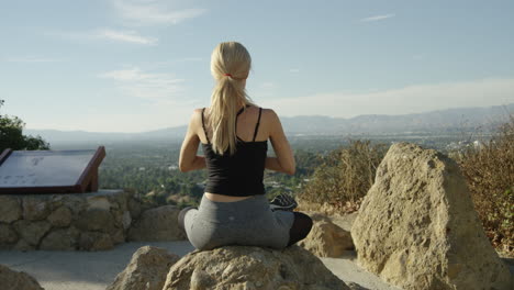 slow motion video of young blonde woman seen from behind sitting on a rock outside in yoga pose with blue sky and beautiful background