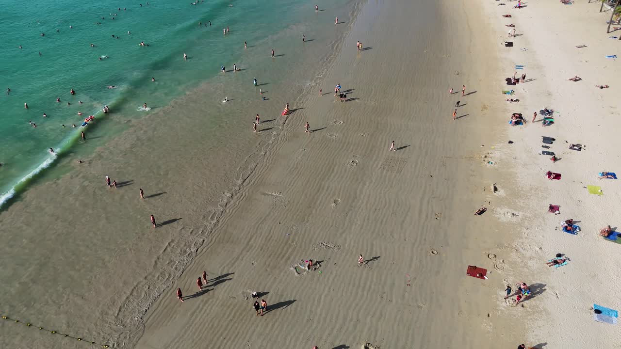 Drone footage captures a vibrant beach scene in Phuket, Thailand, showcasing sunbathers, umbrellas, and turquoise waters under bright sunlight
