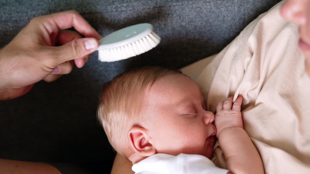Adorable newborn boy lies comfortably in mom's hands. Dad's hand brushing blond child's hair. Close up.