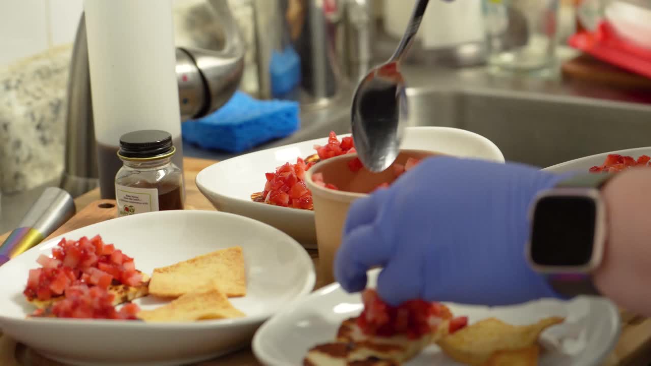 Chef Wearing Blue Rubber Gloves Assembles a Healthy Appetizer Meal by Placing Diced Tomatoes on Pita Bread Chips in the Kitchen using a Spoon and White Bowls or Plates, Food Closeup