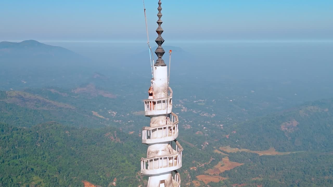 Vertical shot of a woman standing at the top of the iconic Ambuluwawa Tower in Kandy, Sri Lanka. Surrounded by panoramic views of mountains and misty skies.