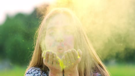 portrait shot of the young beautiful girl blowing paint powder from hands palms and smiling to the camera cheerfully outside