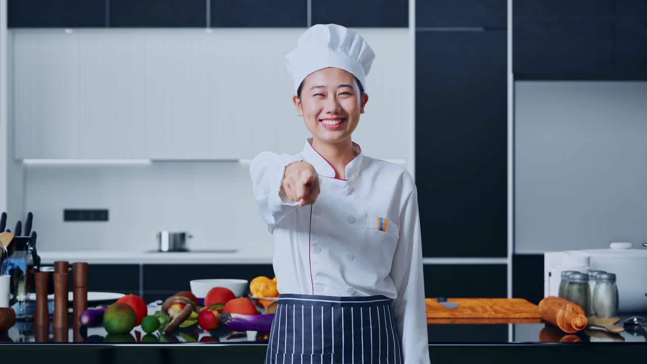 mujer chef asiática sonriendo y tocando su pecho luego señalándote en la cocina de casa