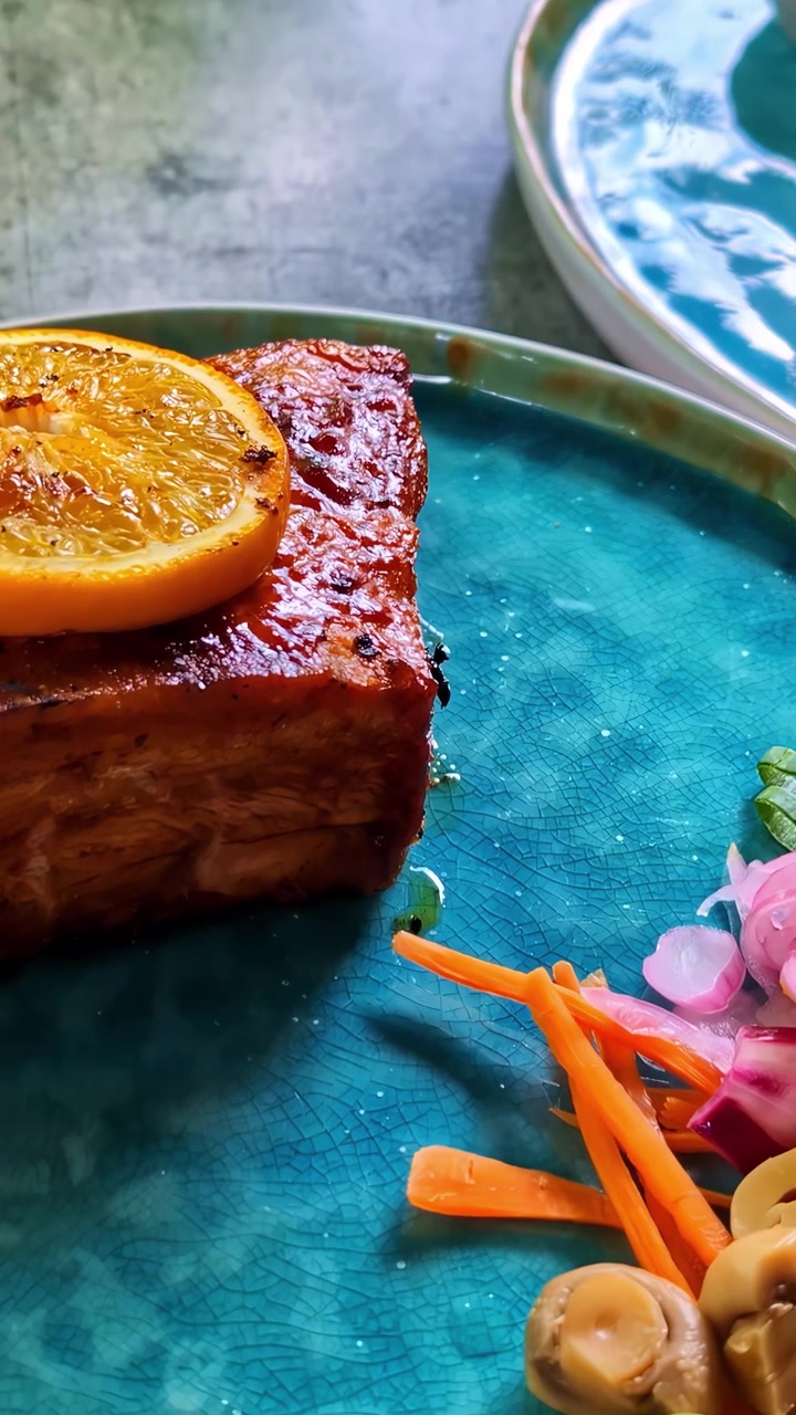 Glazed pork belly served on a blue ceramic dish with orange garnish, pickled vegetables, mushrooms and cucumber ribbon in a restaurant close-up.