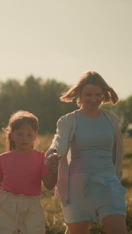 la niña sosteniendo la mano de su madre mira hacia arriba mientras su madre sonríe cálidamente, la tía camina a lo largo sosteniendo una cuerda, haciendo gestos para que el perro se acerque en un día soleado en una vasta tierra de cultivo con árboles lejanos en el fondo