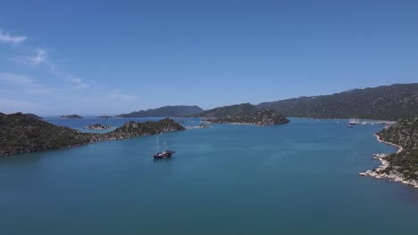 Drone-rises-above-large-ship-with-masts-down-in-Mediterranean-bay-off-the-coast-of-Kas,-Turkey