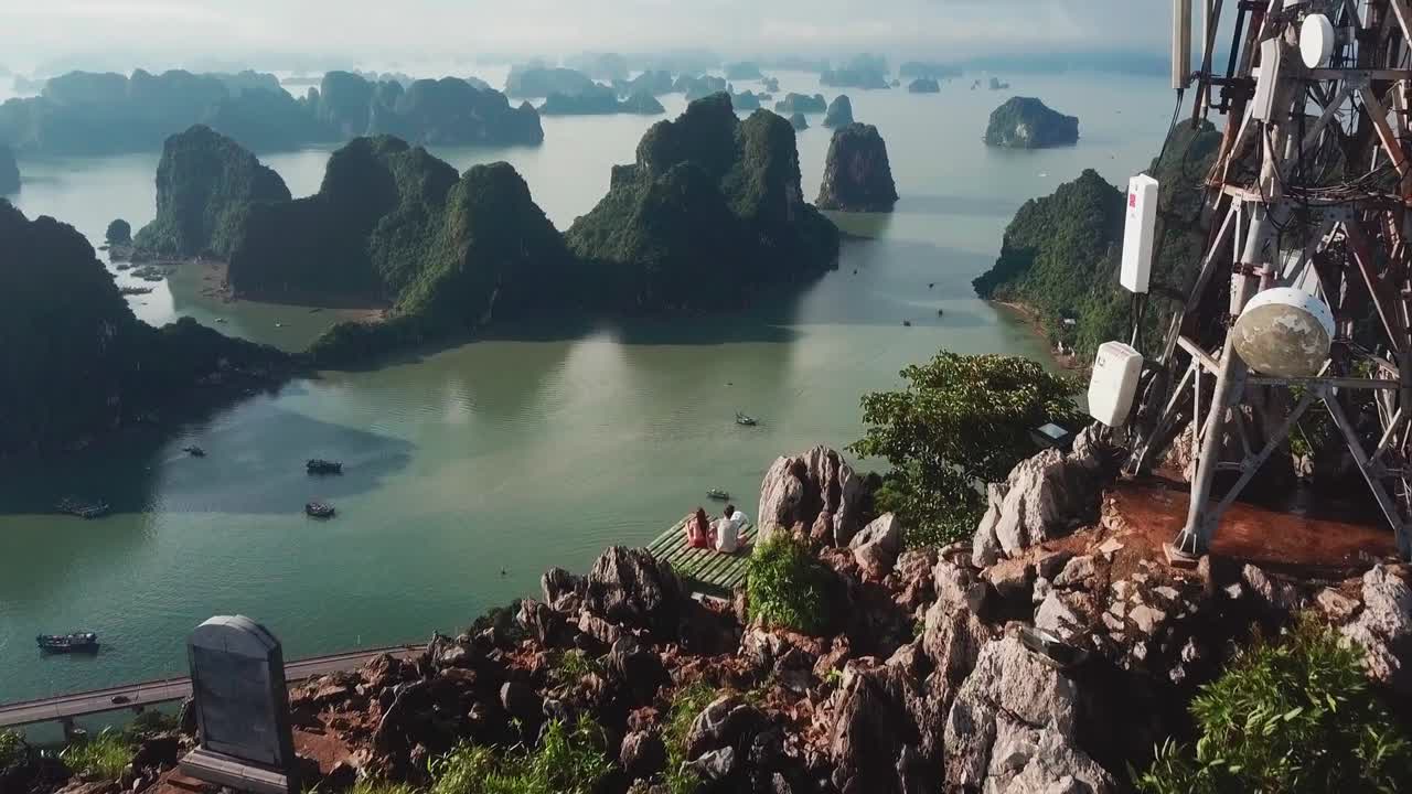 Aerial drone view of Ha Long Bay featuring lush limestone islets rising from calm water, with small boats and distant telecom tower under soft daylight in peaceful natural seascape