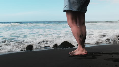 Piernas-De-Mujer,-Pies-En-La-Playa-De-Arena-Negra-En-Hawaii,-Admirando-El-Océano